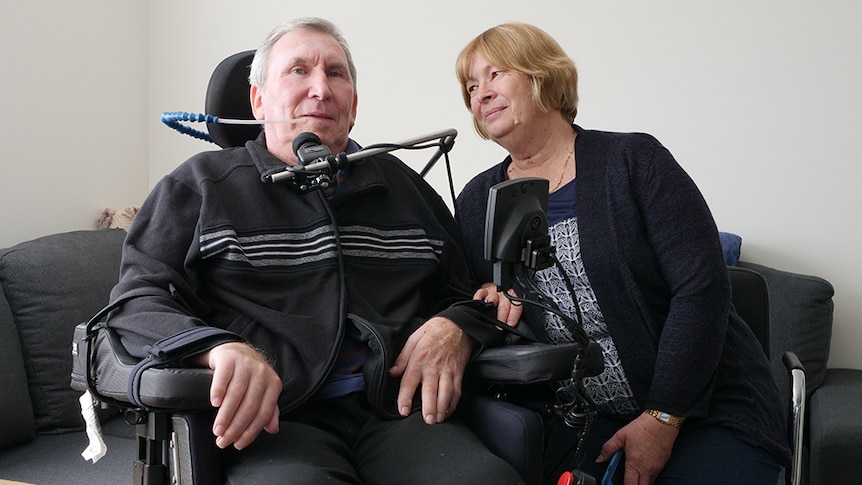 An elderly man in a wheelchair, flanked by his wife.