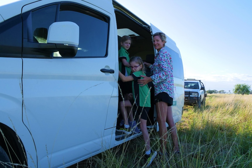 A mother helps two young girls enter a bus on a dirt road