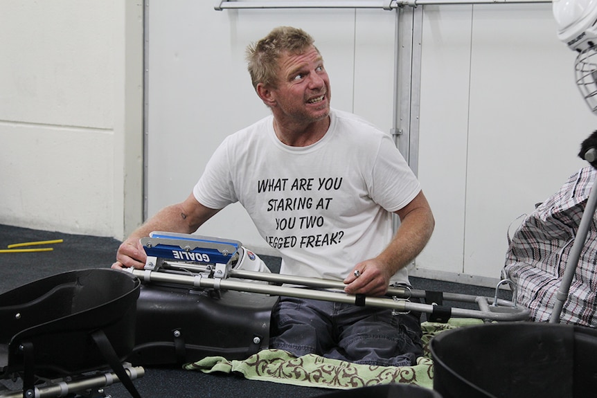 A double amputee sitting on the ground with a plastic and metal sledge hockey sled.