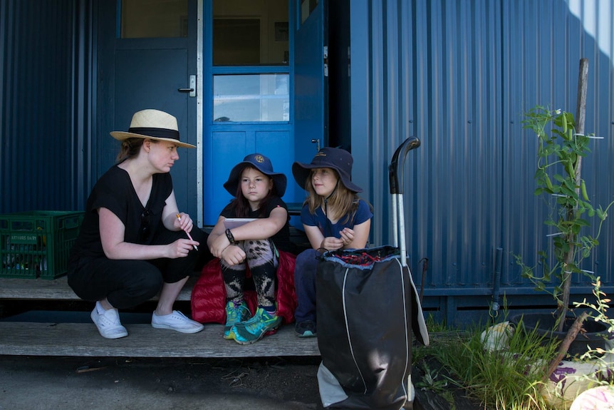 Teacher Anita Harding crouches next to two students sitting on the steps of the school, as they ponder a question she's asked.