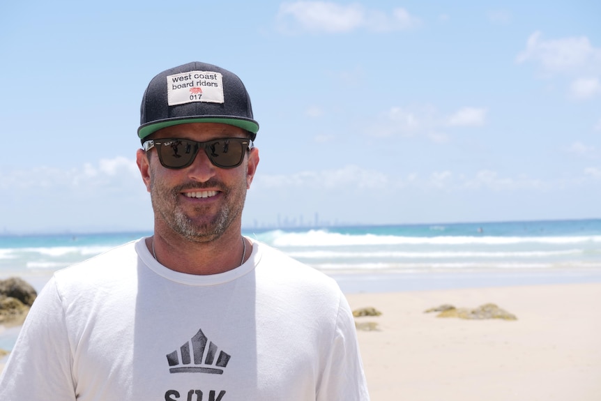 A man in hat and sunglasses stands on a beach smiling.