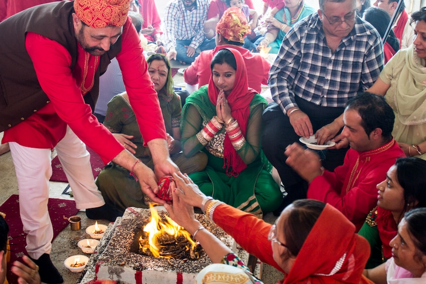 People sit in prayer around a fire, colourful dress, candles and seeds surround fire.