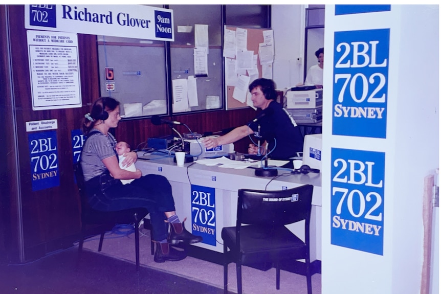 A woman holds a newborn baby as she talks to a man at a desk with 2BL 702 signage. 