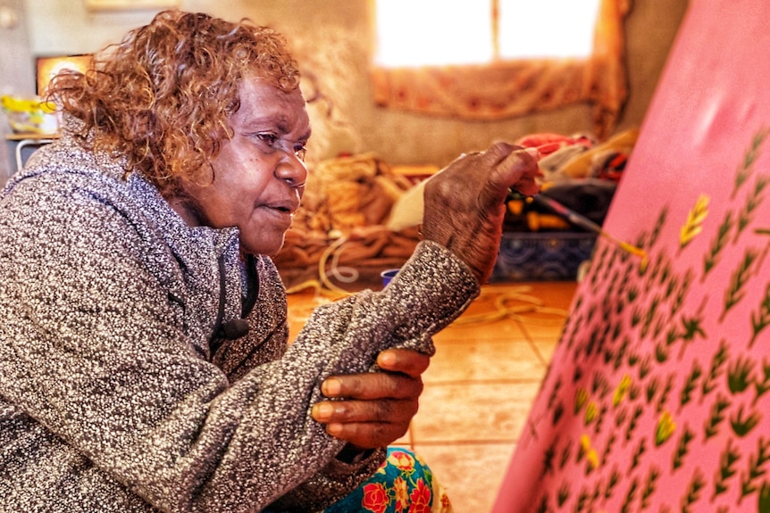 A woman sits on the floor of her home and paints.