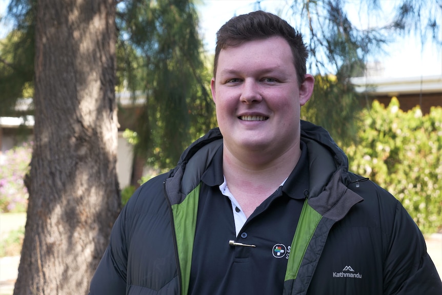 A man in a Kathmandu jacket with a polo shirt underneath smiles at the camera, standing in a leafy garden.