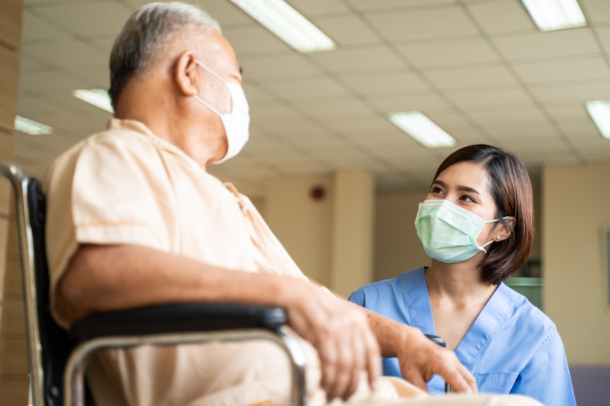 A young nurse wearing a mask and looking at an elderly man in a wheelchair reassuringly.