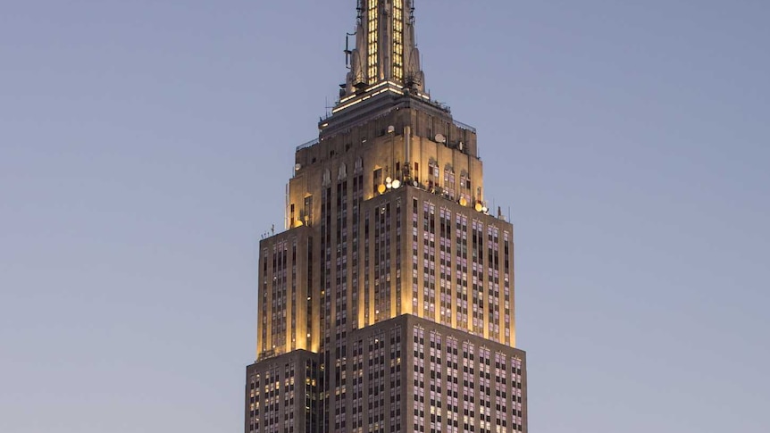 A portrait photo of the Empire State Building in the early evening with the moon in the sky above.