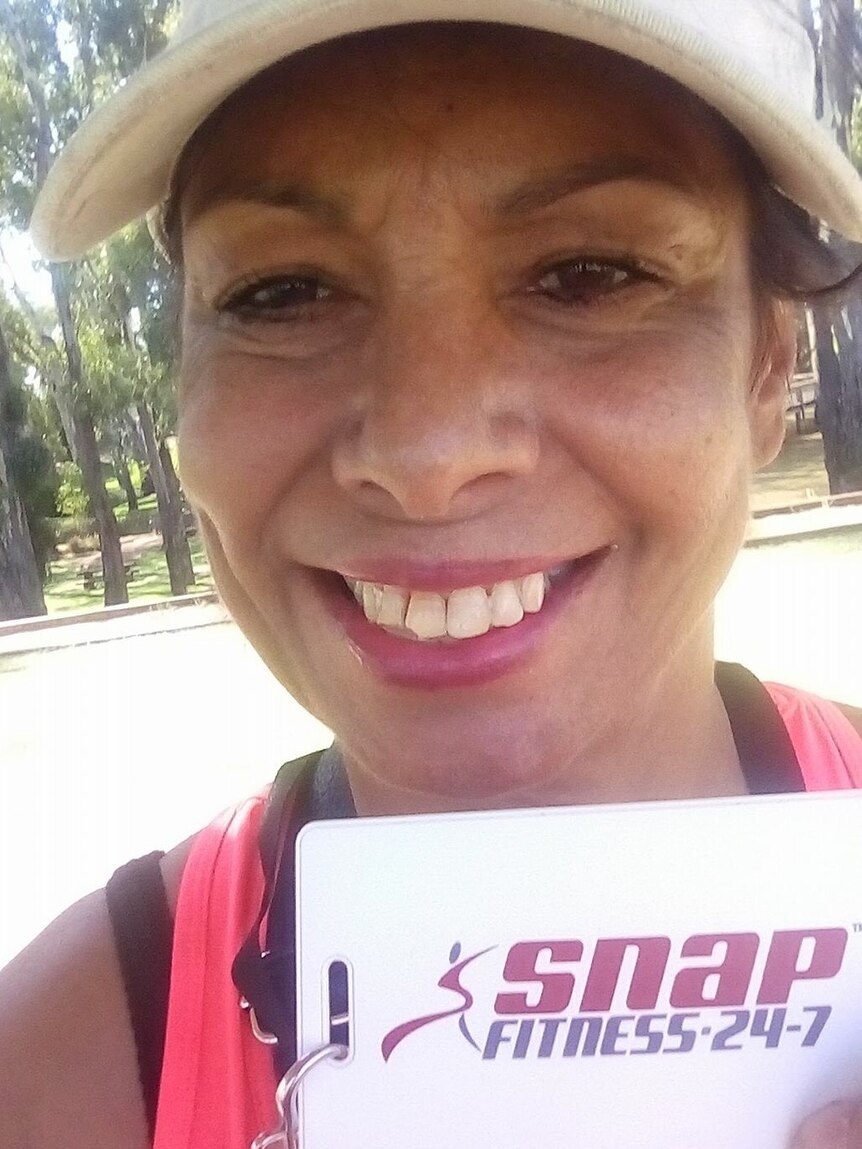Tanya Day, smiling wearing a hat and pink singlet, holds a SNAP fitness card, with trees in the background.