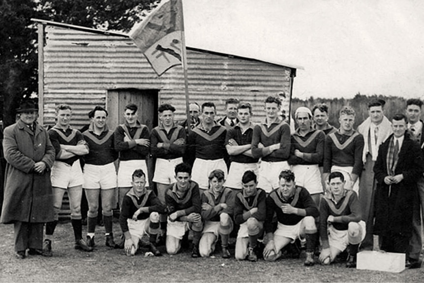 A football team poses outside a rusted corrugated iron change shed, club flag held aloft at the rear.