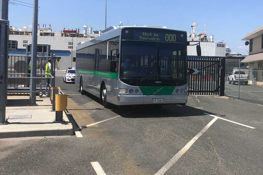 A bus drives out of a secure area of the port, the Al Messilah is in dock behind it.