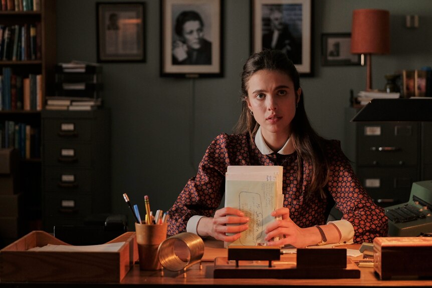 Young woman with long dark brown hair and pale skin, wearing patterned blouse, sitting at desk holding small bundle of letters.