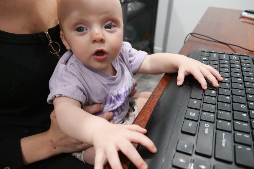 Baby sits on mother's lap with hands on keyboard. 