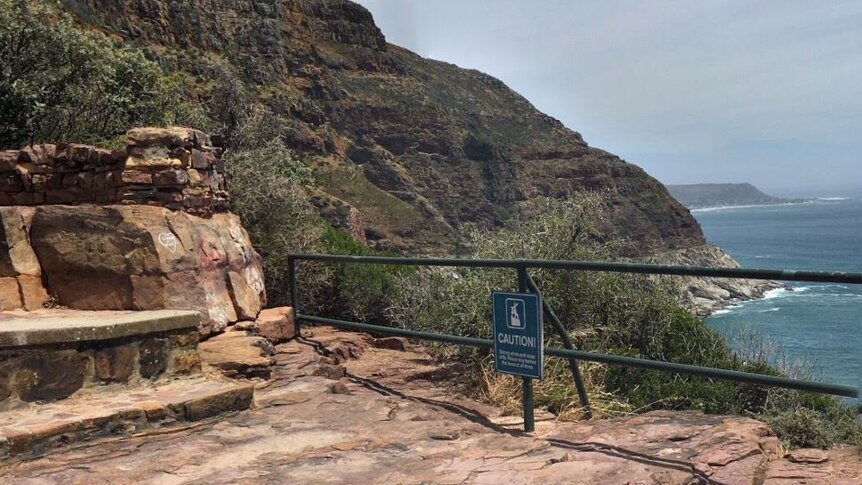A lookout point over a cliff with a guard rail around the edge. There is a sign that reads "caution" stuck to the fence.