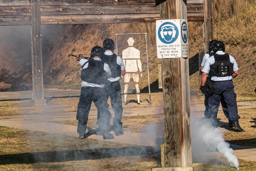 A group of men in protective gear standing in a gun range with a target in front.