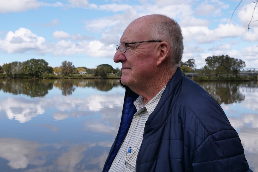 An elderly man looks down at a river, with the sky and clouds reflected in the water.