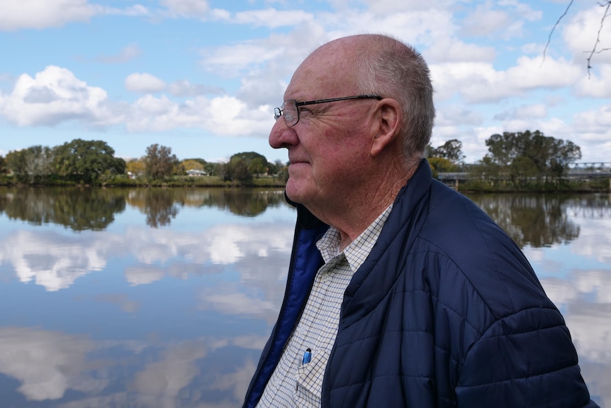 An older man gazes out over a river, with sky and clouds reflected in the water.