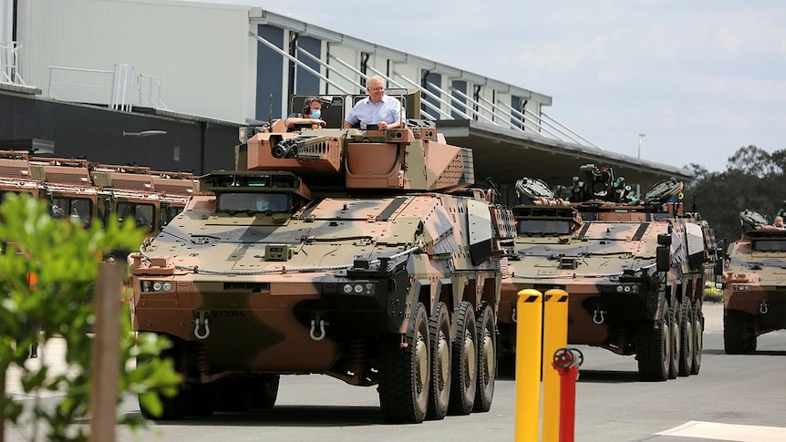 The Prime Minister smiles as he sits atop a tank driving down a street.