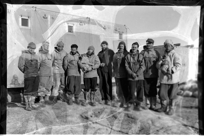 A black and white photo of ten men standing in thick winter clothes in front of a building, two of the men carrying small dogs.