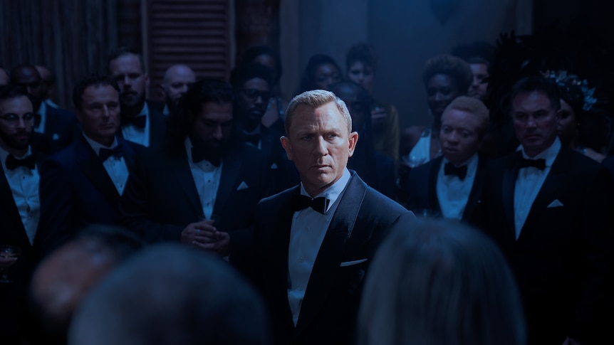 Man with chiselled features and short cropped blonde hair wears a black tuxedo and bow tie in room of elegantly dressed people