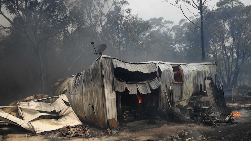 A shed with warped iron walls and collapsed roof amid smoky haze and spot fires.