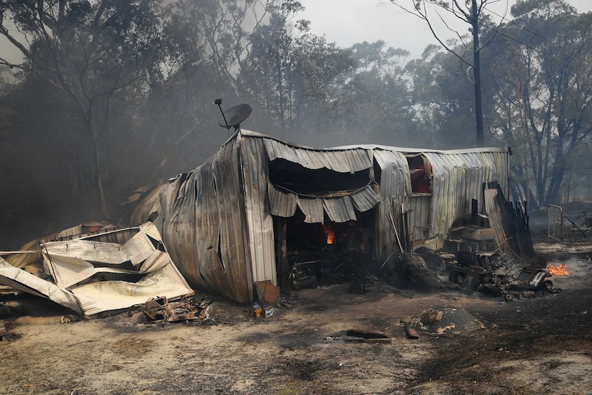 A shed with warped iron walls and collapsed roof amid smoky haze and spot fires.