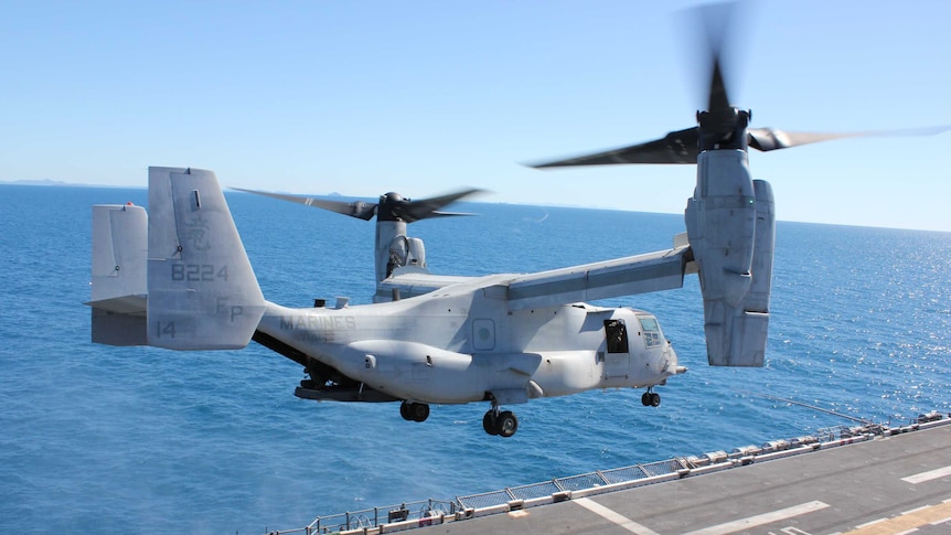 A military helicopter hovers above the deck of an aircraft carrier. The blue sea and sky are visible behind.