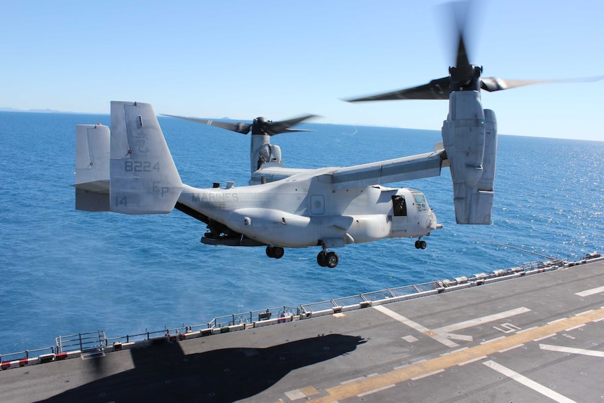 A military helicopter hovers above the deck of an aircraft carrier. The blue sea and sky are visible behind.