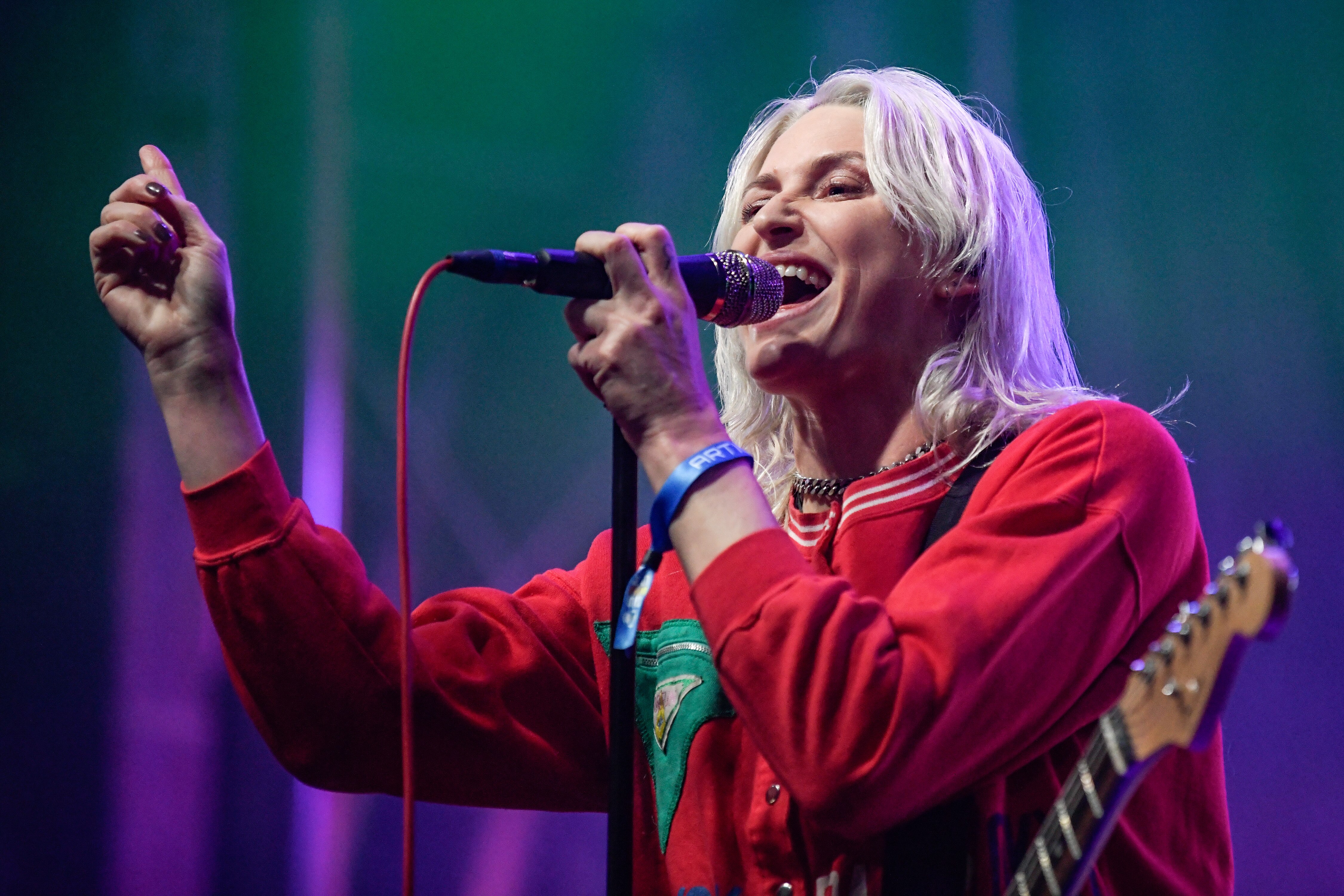 A woman in a red jumper sings into a microphone with an arm raised up.  She has a guitar slung over her shoulder.