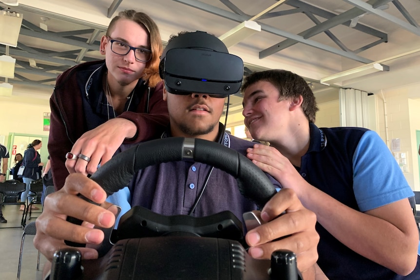A teenage boy using a VR headset and steering wheel while two friends give him advice.