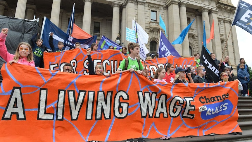 People stand outside a building in Melbourne holding up signs demanding higher wages.