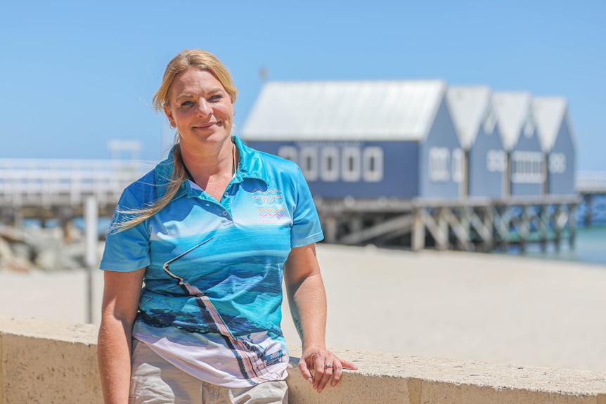 A woman with blonde hair stands at the beach in front of a blue building and jetty.