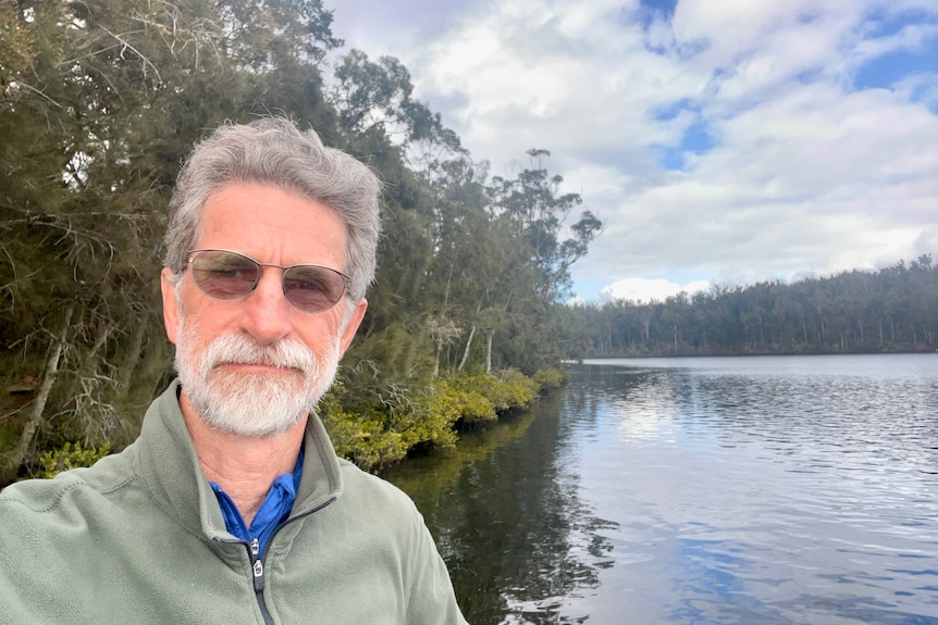 A man wearing glasses and a green jumper takes a selfie with water in the background