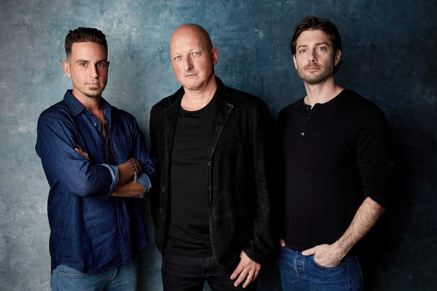 Against a textured aquamarine photo backdrop, three caucasian men pose for the camera.