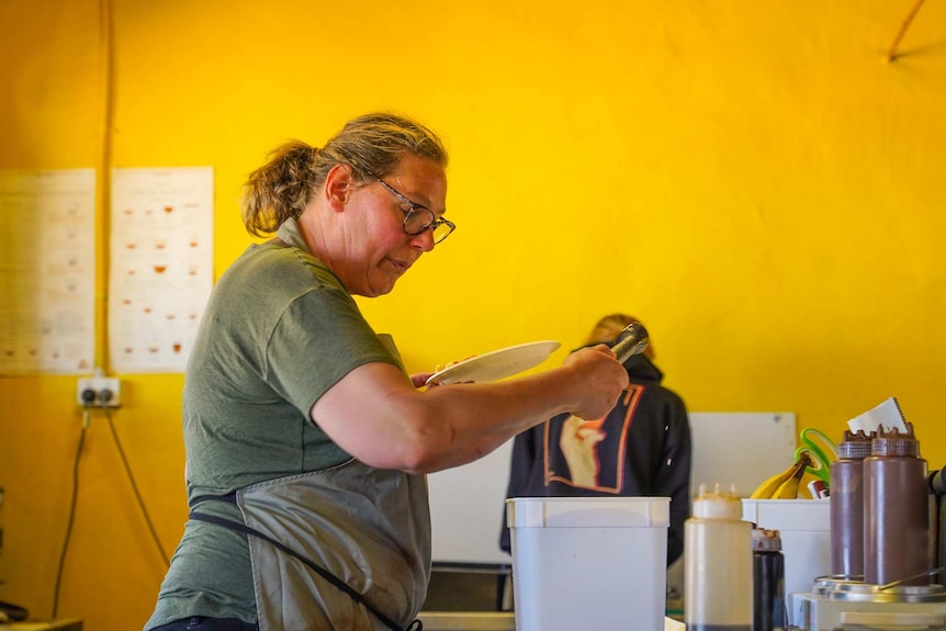 A woman wearing glasses and an apron scoops ice cream from a large bucket in a bright yellow-walled kitchen.