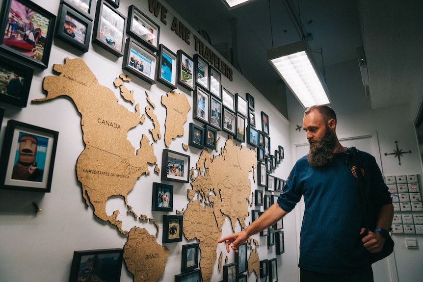 A man with a beard stands and points at a large world map on a wall