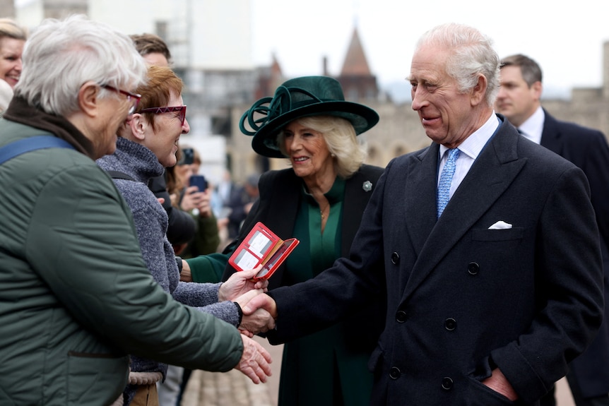 King Charles shakes a woman's hand while walking next to Queen Camilla.