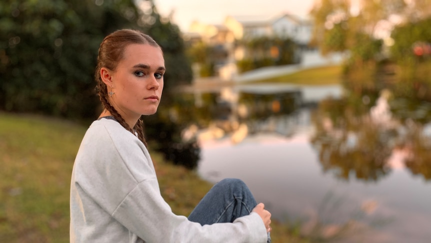 A young woman sits by a body of water, looking serious.