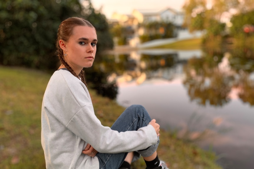 A young woman sits by a body of water, looking serious.