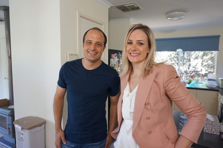 A man with dark hair, wearing a navy t-shirt, standing in a house next to a blonde woman.