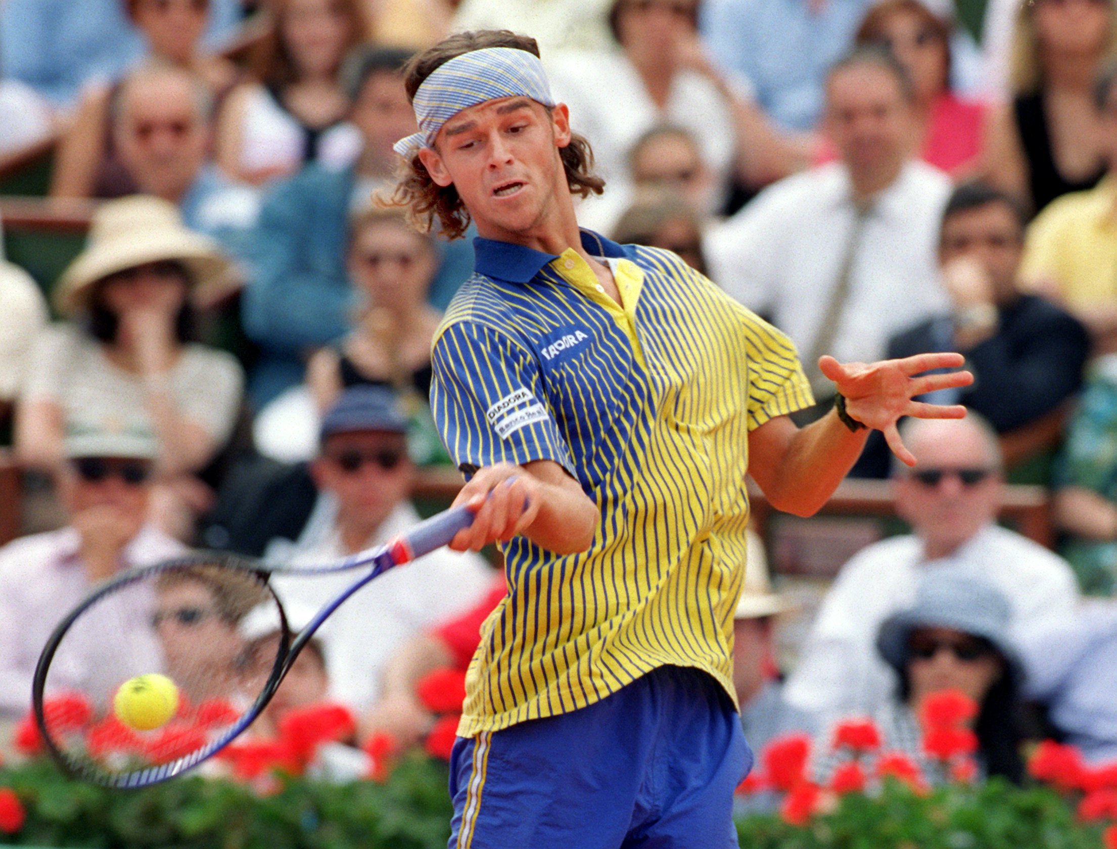 A tennis player wearing a headband stares down at the ball as he stretches to hit a forehand. 
