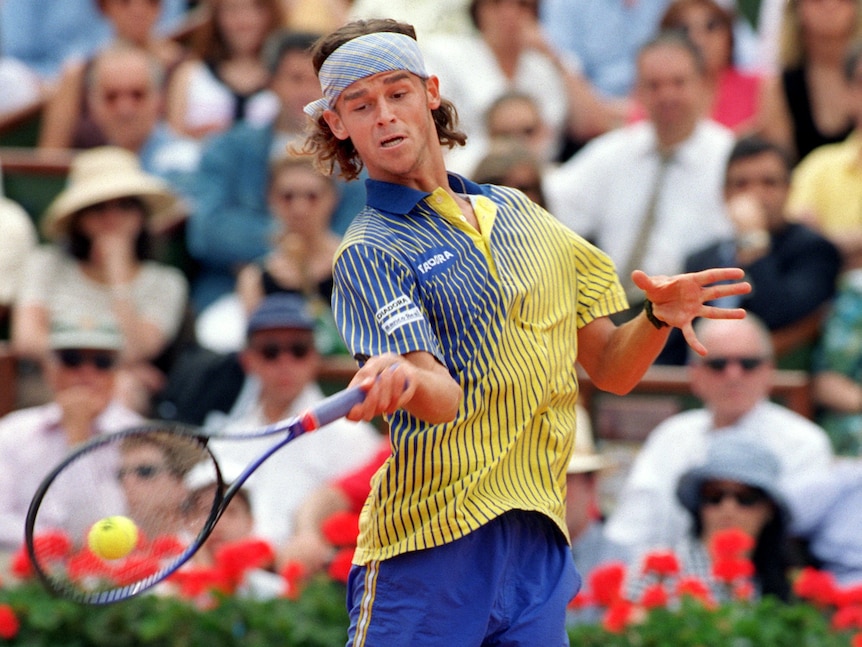 A tennis player wearing a headband stares down at the ball as he stretches to hit a forehand. 