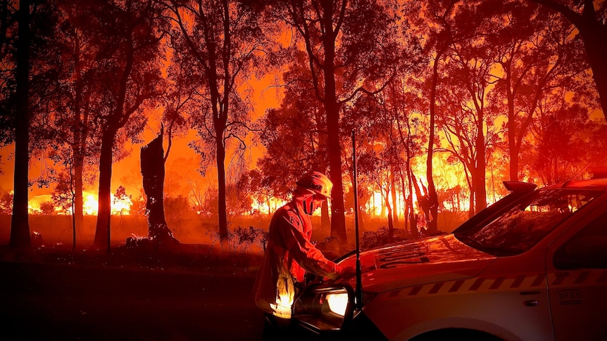 A fierefighter leans on a car as a blaze rages around him at night.