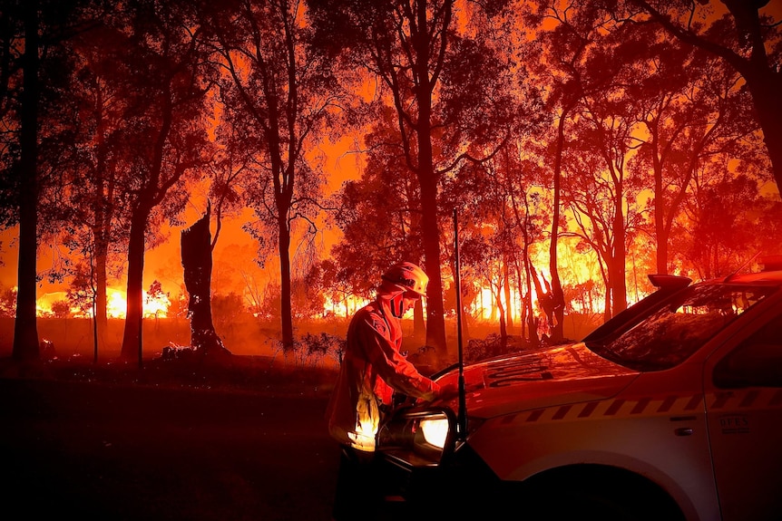 A fierefighter leans on a car as a blaze rages around him at night.