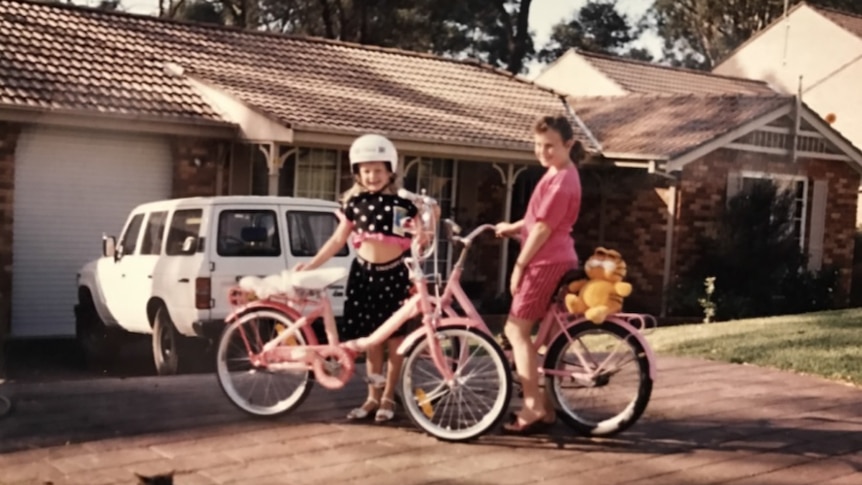 A young Rebel Wilson and another girl with bikes outside a family home.