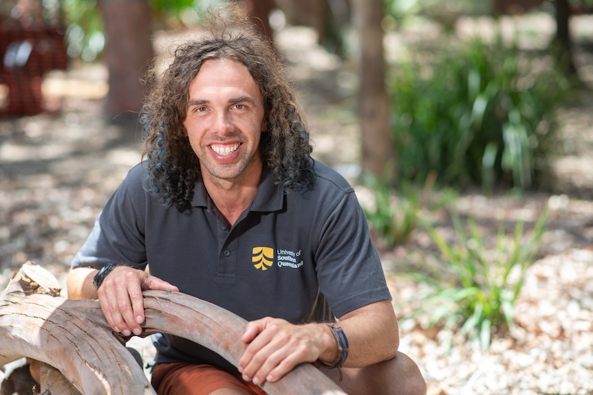 man with curly hair crouches by tree and smiles. 