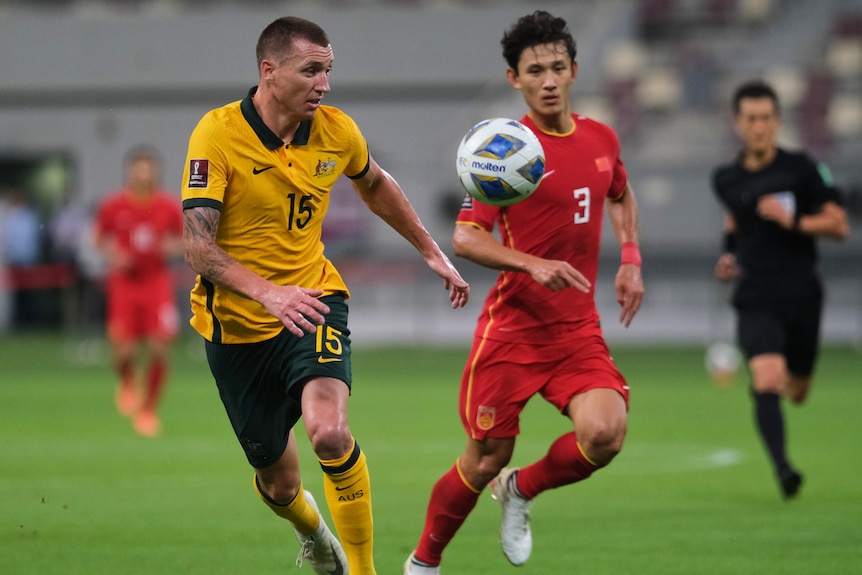 Two soccer players, one wearing yellow and green and another wearing red, chase a ball