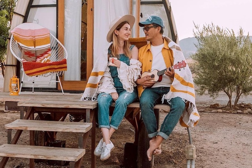 A man and a woman wrapped in a blanket look at each other while sitting on the steps to a cabin in the desert