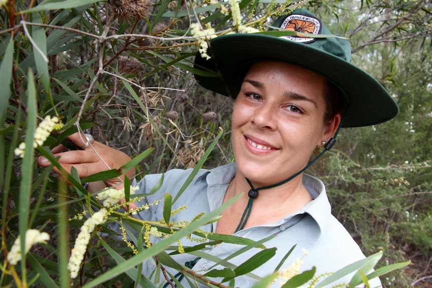 A young woman, Jacinta Rheinberger, wearing a Discovery ranger uniform and hat, inspects a wildflower.