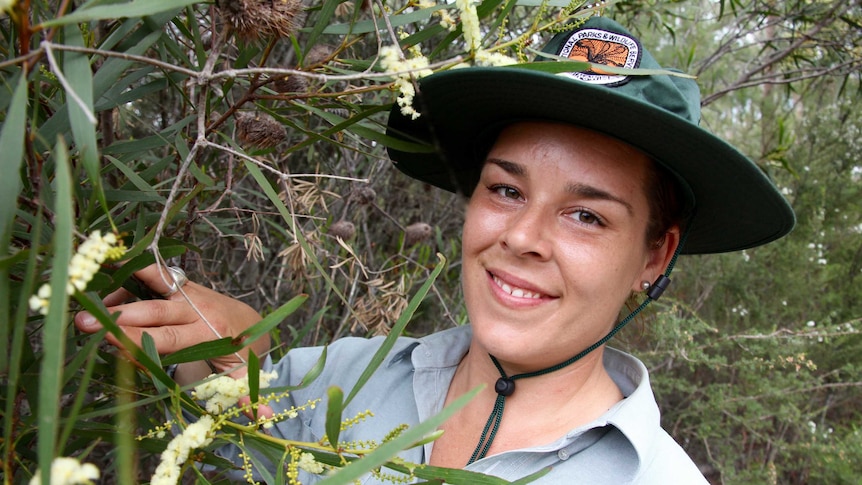 A young woman, Jacinta Rheinberger, wearing a Discovery ranger uniform and hat, inspects a wildflower.