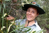 A young woman, Jacinta Rheinberger, wearing a Discovery ranger uniform and hat, inspects a wildflower.